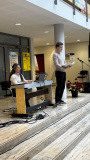 Ein junger Mann steht mit geöffneten Unterlagen neben einem Mikrofon, während eine Frau am E-Piano spielt.