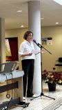 Eine Frau mit weißer Bluse liest an einem Mikrofon von einem Blatt Papier; neben ihr steht ein E-Piano.