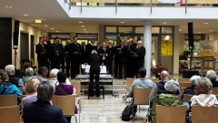 Ein Männerchor singt unter der Leitung einer Dirigentin vor sitzendem Publikum im Foyer des Rathauses.