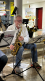 Ein sitzender Musiker mit weißem Hemd spielt Saxofon zwischen zwei Notenständern.