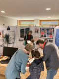 Erwachsene und Kinder beschäftigen sich gemeinsam mit Unterlagen in einem als Büro eingerichteten Raum mit Schreibtisch, Computer und Wandtafeln.