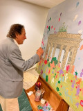 Ein Mann im grauen Sakko ergänzt mit einem Pinsel ein farbenfrohes Gemeinschaftsbild mit dem Brandenburger Tor.
