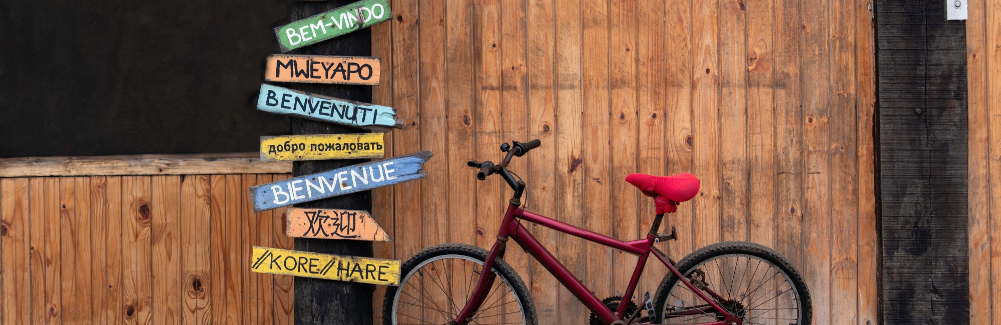 Ein bunter Pfosten mit „Willkommen“ in verschiedenen Sprachen steht vor einer Holzwand, daneben lehnt ein rotes Fahrrad, was eine internationale und gastfreundliche Stimmung erzeugt.