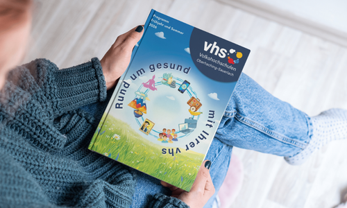 Eine Person hält das Programmheft der vhs Oberhaching mit dem Titel ‚rundum gesund mit ihrer vhs‘ in den Händen.