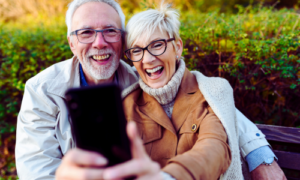 Ein älteres Paar sitzt lachend auf einer Parkbank und macht gemeinsam ein Selfie mit dem Smartphone. Beide tragen moderne Brillen und stilvolle Herbstkleidung. Die Szene strahlt Lebensfreude, digitale Kompetenz und Spaß an moderner Technik aus. Symbolbild für die Smartphone- und Tabletkurse an der vhs Oberhaching, die speziell für Einsteiger und Fortgeschrittene angeboten werden.