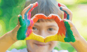 Ein fröhliches Kind zeigt ein Herz aus bunt bemalten Händen – das Bild steht symbolisch für die Kreativität, Lebensfreude und Gemeinschaft, die unser Ferienprogramm für Kinder auszeichnet. Ideal für Familien, die in den Schulferien spannende Freizeitangebote suchen.