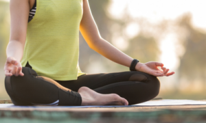 Eine Frau sitzt entspannt im Schneidersitz auf einer Yogamatte und praktiziert eine meditative Haltung. Ihre Hände liegen in einer Mudra-Geste auf den Knien, während der Körper zur Ruhe kommt. Das warme Licht der Abendsonne schafft eine friedvolle Atmosphäre – typisch für eine achtsame Yin-Yoga-Praxis, die auf Entschleunigung, Fasziendehnung und inneres Gleichgewicht abzielt.