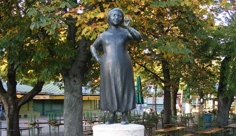 Bronzestatue von Liesl Karlstadt auf einem Brunnen am Viktualienmarkt in München, umgeben von Bäumen.
