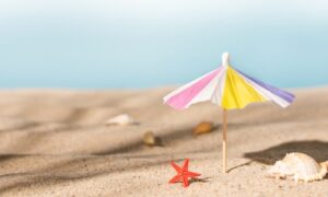 Ein kleiner, bunter Papier-Sonnenschirm steckt im hellen Sand eines Strandes. Daneben liegen eine rote Seesternfigur und einige Muscheln. Im Hintergrund verläuft der Horizont in weichen Blautönen – es entsteht eine ruhige Sommerstimmung. Unsere Geschäftsstelle bleibt im August für den Publikumsverkehr geschlossen. Per E-Mail sind wir weiterhin erreichbar und freuen uns auf Ihre Nachricht.
