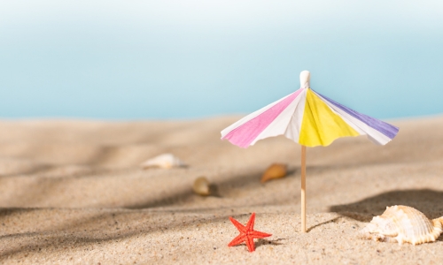 Ein kleiner, bunter Papier-Sonnenschirm steckt im hellen Sand eines Strandes. Daneben liegen eine rote Seesternfigur und einige Muscheln. Im Hintergrund verläuft der Horizont in weichen Blautönen – es entsteht eine ruhige Sommerstimmung. Unsere Geschäftsstelle bleibt im August für den Publikumsverkehr geschlossen. Per E-Mail sind wir weiterhin erreichbar und freuen uns auf Ihre Nachricht.