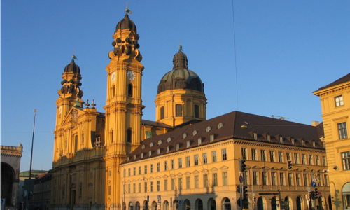 Die Aufnahme zeigt die prächtige Fassade der Theatinerkirche St. Kajetan in München, ein herausragendes Beispiel des süddeutschen Barocks. Im Vordergrund erheben sich die beiden markanten Zwiebeltürme, deren gelblich-ockerfarbener Putz im Sonnenlicht warm leuchtet. Dazwischen thront die reich verzierte Hauptfassade mit ihrer geschwungenen Giebelstruktur und kunstvollen Stuckelementen. Der Blickwinkel der Fotografie lässt die imposante Höhe der Kirche deutlich erkennen. Im unteren Bildbereich sind Teile des geschäftigen Odeonsplatzes zu sehen, was das Zusammenspiel zwischen historischer Architektur und urbanem Leben betont. Der klare Himmel im Hintergrund unterstreicht die monumentale Erscheinung dieses Münchner Wahrzeichens.
