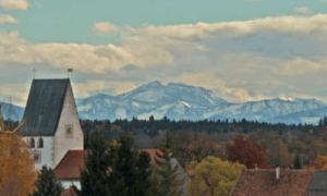 Das Bild zeigt die Pfarrkirche St. Stephan in Oberhaching mit ihrem steilen Satteldach vor herbstlich gefärbten Bäumen und den Dächern des Ortes. Im Hintergrund erhebt sich eindrucksvoll die schneebedeckte Alpenkette unter einem teils bewölkten Himmel. Die Aufnahme verbindet dörfliche Idylle mit majestätischer Fernsicht.