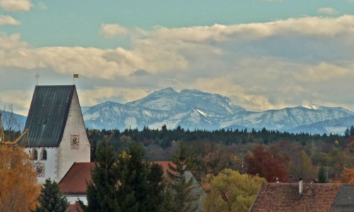 Das Bild zeigt die Pfarrkirche St. Stephan in Oberhaching mit ihrem steilen Satteldach vor herbstlich gefärbten Bäumen und den Dächern des Ortes. Im Hintergrund erhebt sich eindrucksvoll die schneebedeckte Alpenkette unter einem teils bewölkten Himmel. Die Aufnahme verbindet dörfliche Idylle mit majestätischer Fernsicht.