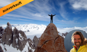 Thomas Huber steht mit ausgebreiteten Armen auf einem Berggipfel umgeben von schneebedeckten Felsen