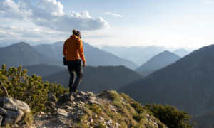 Eine Person mit orangefarbener Jacke steht auf einem felsigen Berggrat mit Blick auf ein weites Alpenpanorama.