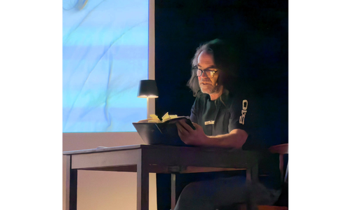 Thomas Huber sitzt bei seinem Multivisionsvortrag im Bürgersaal Oberhaching an einem Tisch und liest bei gedämpftem Licht aus einem Notizbuch. Neben ihm steht eine kleine Lampe, im Hintergrund ist eine Projektion zu sehen.