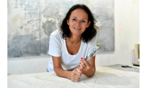 Eine Person ist in einem Atelier vor einer Leinwand über einen Tisch gebeugt und hält mehrere Pinsel in der Hand.