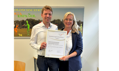 Dr. Felix Bokelmann und Johanna Weizer-Hesky halten gemeinsam eine gerahmte Anerkennungsurkunde für Excellence Level 2 in einem VHS-Kursraum.